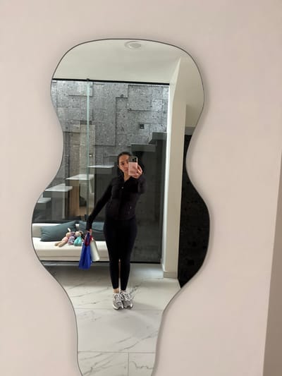 A woman wearing a black athletic outfit takes a mirror selfie. The mirror has an unusual, guitar-like shape and is hanging on a white wall. The reflection shows a hallway with gray tiled stairs, a glass railing, and a white marble floor. The woman holds shopping bags and a stuffed animal.