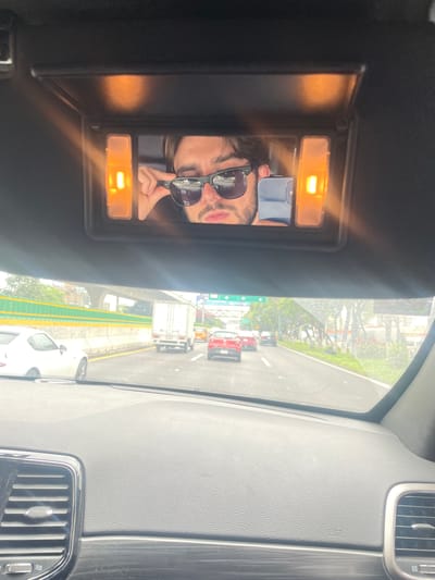 The image shows a person reflected in a car's vanity mirror, holding a phone to take a selfie. The person is wearing sunglasses. The car is on a highway in Tlalnepantla, Mexico, with traffic visible through the windshield. The highway is lined with trees and buildings. The weather appears to be overcast.