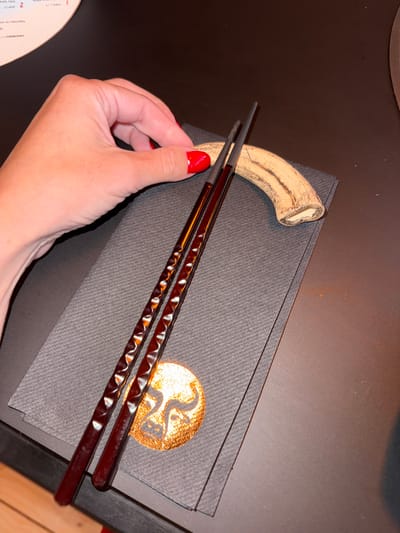 The image shows a pair of brown chopsticks resting on a dark gray textured surface. A hand with red nail polish is holding one end of the chopsticks. A small, light-colored, curved object is next to the chopsticks on the upper left. A gold-colored logo is visible on the dark gray surface. A circular object with text is partially visible in the lower left. The setting appears to be a restaurant table with a dark surface. The city and country of Cape Town, South Africa are not discernable from this single image.