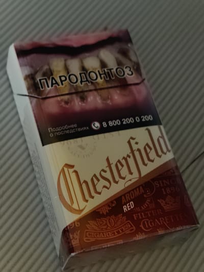 This media file shows a close-up, angled view of a pack of Chesterfield cigarettes. The pack is predominantly white with red and brown accents. The top of the pack features a graphic image of unhealthy teeth and gums, with the Russian word "ПАРОДОНТОЗ" (periodontitis) prominently displayed in large, bold, white lettering against a dark background. Below this, smaller Russian text reads "Подробнее о последствиях" (More about the consequences), alongside a phone number "8 800 200 0 200" and a phone icon.
The lower portion of the pack displays the brand name "Chesterfield" in large, stylized, golden-brown lettering. Below this, a red band contains further details. The words "AROMA", "RED", and "FILTER" are visible, along with "SINCE 1896". Decorative elements, including crowns and flourishes, are present, as well as the text "20 CIGARETTES". The pack appears to be wrapped in cellophane, reflecting the light. The background consists of a textured surface with diagonal lines, suggesting a table or countertop. There is no indication of time of day, weather, or specific location cues beyond the Russian text on the packaging. The overall presentation is that of a product advertisement, with a health warning integrated into the design.