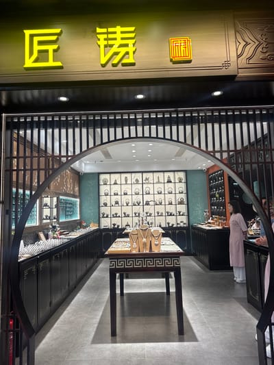 The image depicts the entrance and partial interior of a retail store, presumed to be located in Qingyang District, China. Above the dark wooden entrance facade, a neon yellow-green sign displays three Chinese characters: "匠" (jiàng, craftsman), "铸" (zhù, to cast/forge), and a stylized square character resembling a seal script "坊" (fāng, workshop/store), together reading "匠铸坊" (Craftsman's Forge/Casting Workshop).

The entrance features a dark wooden archway framed by vertical wooden slats above. Inside, the store utilizes a dark wood aesthetic with intricate patterns. On the left interior wall, a brown panel displays white floral patterns, resembling cherry blossoms. The right interior wall is covered in a teal-colored wallpaper with subtle light patterns. The ceiling features multiple recessed spotlights providing bright illumination.

In the center of the foreground, a dark wooden display table with white Greek key patterns on its side supports multiple jewelry busts showcasing ornate silver-toned necklaces. Along the left side of the store, a long, dark wooden counter with cabinets underneath is topped with numerous small, shiny silver items, possibly rings or bracelets. Further back on the left, additional illuminated display cases are visible.

On the right side of the store, a large wall-mounted display unit with square compartments showcases a collection of silver-toned metalware, predominantly small teapots or kettles, alongside a few figurines including a horse and rider, and a Buddha statue. Adjacent to this, a narrower vertical display holds multiple small white cups. A woman in a light pink tunic dress stands to the right, facing towards the left side of the store. Her hands are clasped in front of her. Partially visible on the far right are other individuals in light-colored clothing. The floor is covered in light gray tiles.