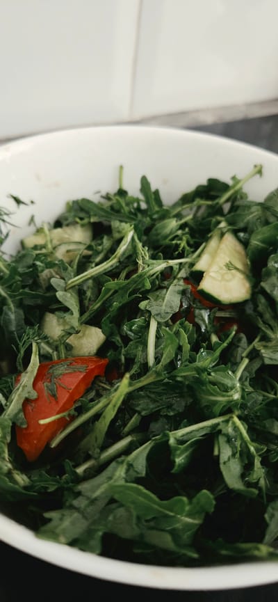 A close-up, overhead shot shows a white bowl filled with a vibrant green salad. The salad is composed of arugula leaves, thin slices of cucumber, and chunks of red tomato. A hint of dill is also visible, adding a delicate touch to the mix. The background is blurred, but the white of the bowl and the kitchen tile are discernible. The image does not depict any specific location in Lipetsk, Russia.