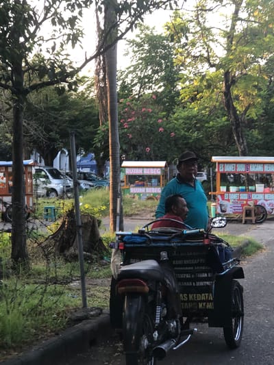 This is a sideways shot of two people on a motorcycle with a sidecar. The person in front is wearing a blue shirt and a dark cap, and appears to be smoking. Behind them, a child in a red shirt is peeking out from the sidecar. The motorcycle has a sign on the sidecar with text in Indonesian. The setting appears to be a roadside with trees and some foliage. There are also some stalls in the background, one with the text "ES DAWET SUSU KACANG MERAH" and "RUJAK BEBEG", suggesting a food and drink vendor. Another stall has "DURIAN", "MANOVA", and "ALPUKAT". The image looks like it was taken during the day, possibly late afternoon, given the soft lighting. The ground is paved, and there is a barbed wire fence in the foreground. The overall scene suggests a casual roadside stop in a tropical or subtropical region, likely in Indonesia given the language on the signs.
