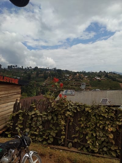 The scene depicts a panoramic view of a hilly, vegetated landscape in Bwasinge, Democratic Republic of the Congo, under a partly cloudy sky. In the immediate foreground, a black motorcycle with a silver front wheel is parked on a dirt surface at the bottom left. To its right, a rustic wooden fence constructed from vertical slats is heavily covered by a dense vine with numerous green leaves. Red flowers are visible above the fence. Behind the fence and below the horizon, a corrugated metal roof is present, featuring a small white satellite dish and a square solar panel. To the far left, a light-colored wooden structure or building, composed of horizontal planks, displays partial text that reads "PALESTINE." The midground and background consist of rolling hills densely covered in green vegetation, interspersed with numerous scattered houses and structures, indicating a populated rural or semi-urban area. No individuals are visible in the image.