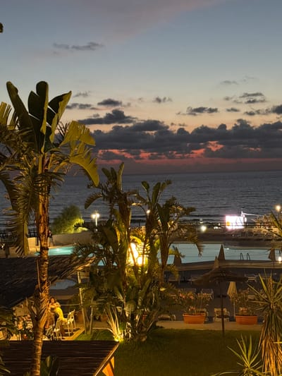 The image depicts an outdoor scene at dusk, likely at a resort or beachfront establishment in Capaci, Italy. In the foreground, large palm trees with broad leaves are visible, some illuminated by artificial lighting. Beyond the trees, a swimming pool with clear blue water is partially seen. Several lounge chairs and umbrellas are arranged around the pool area, some of which are occupied or being used.
The middle ground of the image features a view of the sea, which is dark and calm under the twilight sky. The sky itself is a gradient of colors, transitioning from a pale blue at the top to hues of pink and orange near the horizon, with scattered dark clouds. A bright, illuminated rectangular screen is visible in the distance over the water, possibly a projection or a large LED display.
In the foreground and mid-ground, several people are present. On the left, two individuals are seated at a table with chairs, appearing to be dining or socializing. Another person is walking near the edge of the pool. Further in the background, near the beach area, more people and the outlines of structures can be discerned. The overall atmosphere is tranquil and relaxed, suggesting an evening setting at a leisure destination.