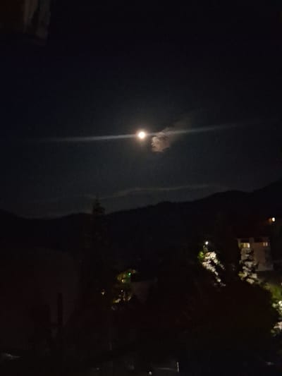 This is a nighttime photograph taken in Smolyan, Bulgaria. The scene is predominantly dark, with a large, vaguely illuminated vertical shape dominating the left side of the frame. This shape could be a building or a natural formation, and a bright, circular light source, possibly the moon or a streetlamp, is visible against it, casting a faint glow and a vertical streak of light upwards.

In the upper right portion of the image, there are some faint lights from what appears to be buildings and trees. The lights suggest an urban or semi-urban setting, with some illumination from windows or outdoor lighting. The darkness indicates that it is late at night or very early morning. No specific activity or event is clearly depicted due to the low light conditions and the abstract nature of the illuminated shapes. The overall mood is subdued and somewhat mysterious due to the limited visibility. There is no visible text in the image.