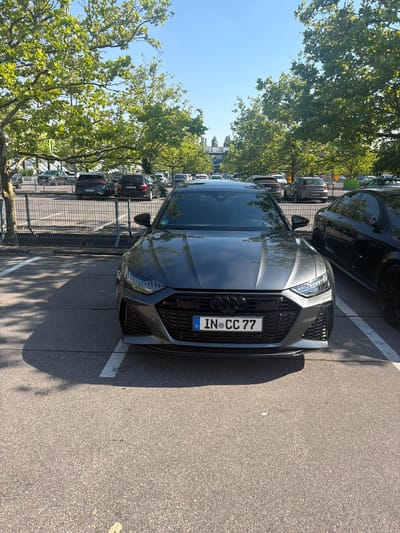 A dark grey Audi RS6 sedan is parked in an outdoor asphalt parking lot. The car is facing the camera and is the primary subject. The license plate reads "IN CC 77". The car is positioned between two white parking lines, with a shadow from a tree falling across the front left portion of the vehicle. In the background, there is a metal fence with parked cars visible behind it. Further in the distance, trees line the horizon under a clear blue sky. The overall environment appears to be a sunny day in a parking lot. Based on the license plate and the company logo on a building in the background, the location is likely the Audi AG headquarters in Ingolstadt, Germany. There are no people visible in the image.
