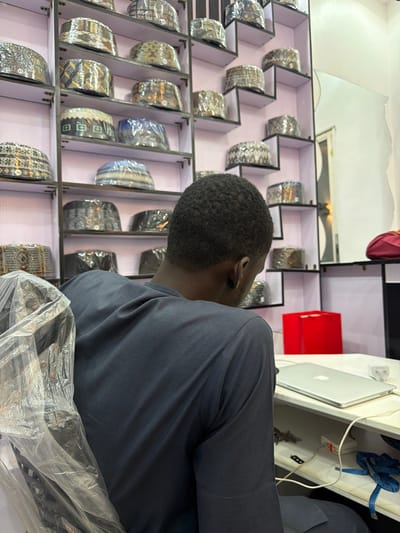The image displays an indoor scene in Gandu Albasa, Nigeria. An adult individual is seated with their back to the camera, wearing a dark grey long-sleeved garment. Their short, dark, kinky hair is visible. The person is positioned in a chair, partially covered on the left by a clear plastic bag. To the individual's right, a light-colored desk holds a closed silver-colored laptop, a red rectangular box, a white power strip with multiple connected wires, a white charging brick, and a blue coiled measuring tape.
The background consists of a wall lined with numerous shelves, predominantly dark-framed with light backings. These shelves display a large collection of traditional caps, each individually wrapped in clear plastic. The caps exhibit various patterns and colors. On the far right wall, a vertical rectangular mirror is present, reflecting a soft, indistinct light source. Below the mirror, a red fabric item rests on a shelf or surface.