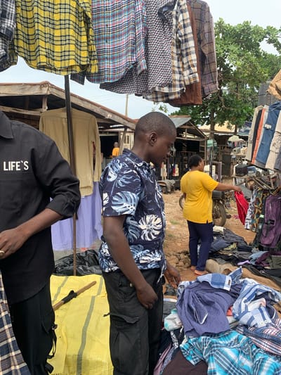 The image depicts an outdoor market scene in Nyanya, Nigeria. The foreground features two individuals. One person, a man, is wearing a dark shirt with a blue and white floral pattern and dark pants. He is partially visible, with his head and torso to the left, and his hand resting on his leg. The other visible person is a woman in a bright yellow t-shirt and dark leggings. She is holding up a red piece of fabric, possibly a garment.
The background is filled with piles of clothing, suggesting a marketplace where goods are sold. Various patterned shirts, including plaid and checkered designs in multiple colors, are stacked and displayed. Some of the clothing appears to be hanging on makeshift structures. There are also hints of other market stalls and possibly some motorized vehicles, like a motorcycle. The overall setting appears to be rustic, with wooden structures and uneven ground.
The lighting suggests it is daytime, and the weather is likely warm, given the outdoor setting and the attire of the people. The atmosphere seems to be that of a busy commercial area, with a focus on the sale of textiles and clothing. The image captures a snapshot of everyday commerce in a Nigerian market.
There is some visible text on the black shirt worn by the person on the bottom, reading "LIFE'S". The overall impression is that of a vibrant and active marketplace.