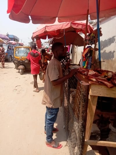 The image depicts an outdoor market scene in Ogidi, Nigeria, under daylight conditions. In the foreground right, a market stall displays raw meat cuts, including ribs and other portions, on a wooden surface. A dark-complexioned man with a short beard, wearing a light brown striped short-sleeved top, blue jeans, and red-soled sandals, stands facing the stall, leaning with his hands near the counter. Below the counter, behind a metal grate, the head of a horned animal, likely a goat or ram, is partially visible. A large red market umbrella provides shade over this area. To the left, a second dark-complexioned man in a red short-sleeved top and flip-flops walks away from the viewer. Behind him is a yellow Keke Napep (motorized tricycle taxi). A blue van is partially visible further in the background, amidst more market activity, additional vendors, and umbrellas. The ground is composed of packed dirt. A light-colored building wall forms the background on the right, exhibiting shadow patterns.