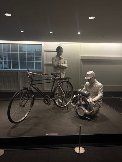 This image depicts a diorama inside a museum, likely in Nagoya, Japan, showcasing historical industrial or technological development. Two life-sized mannequins, sculpted in a monochromatic, realistic style, are the central subjects.

On the left, a mannequin stands upright, dressed in a simple uniform with rolled-up sleeves, wearing glasses, and with his chin resting on his hand in a contemplative pose. Beside him is a vintage bicycle.

On the right, a second mannequin, also in a uniform and wearing a cap, is kneeling and appears to be working on a small engine or mechanical device. The engine is mounted on a stand and features visible pipes and a circular component.

The mannequins and the bicycle are placed on a dark, elevated platform. Behind them, a stylized backdrop features a large window with a grid pattern, revealing an image of industrial buildings or factory structures. To the right of the window, a textured wall with vertical ridges is visible, and further to the right, a plain light-colored wall.

The lighting in the scene is subdued, emanating from spotlights on the ceiling, creating distinct shadows on the platform. A thin rope barrier runs in front of the display, and a small sign with Japanese text and an icon is attached to the edge of the platform. The overall atmosphere is one of historical preservation and exhibition.