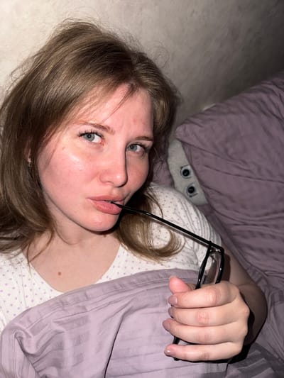 A young woman lies in bed, holding black-rimmed glasses between her fingers and mouth. Her head is tilted towards the camera, revealing light brown hair, blue eyes, and fair skin. The bed is covered with lavender-colored bedding, including a pillow directly behind her head. Partially visible behind the pillow is a gray stuffed animal with prominent eyes. The woman wears a white top with lavender polka dots. The scene appears to be indoors, possibly a bedroom. No information indicates this image was taken in Gornyi (Dvurechenskoe S/P), Russia.