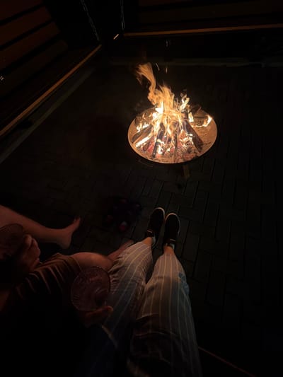 The image depicts a nighttime gathering in Kamennoye, Russia. A circular fire pit filled with burning wood is prominently featured, casting light onto a brick-paved ground. Two individuals are visible: one is wearing striped pants and black shoes, the other is barefoot and holding a glass. A dark object, possibly a bag or container, is placed near the fire pit. The surrounding environment appears to be a dark outdoor space, with a glimpse of a wooden structure on the left.