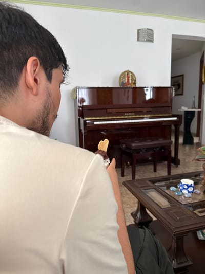 The scene depicts an interior residential space within the city of AMCO, Area Metropolitana Centro Occidente, Colombia. A male individual is visible from the back and right side, wearing a white t-shirt. He has dark hair and stubble along his jawline. He holds a transparent plastic container containing a dark dessert, surmounted by a heart-shaped wafer. In the background, a brown Kawai upright piano is positioned against a white wall, accompanied by a matching brown piano bench. A golden-framed decorative object is mounted on the wall directly above the piano. To the right of the piano, an open doorway reveals an adjacent room. A wall-mounted light fixture is visible on the upper section of the white wall. In the right foreground, a dark wooden coffee table with a glass top holds a small blue and white patterned ceramic cup and multiple smooth, painted stones. A potted plant is partially visible on the table's right edge.