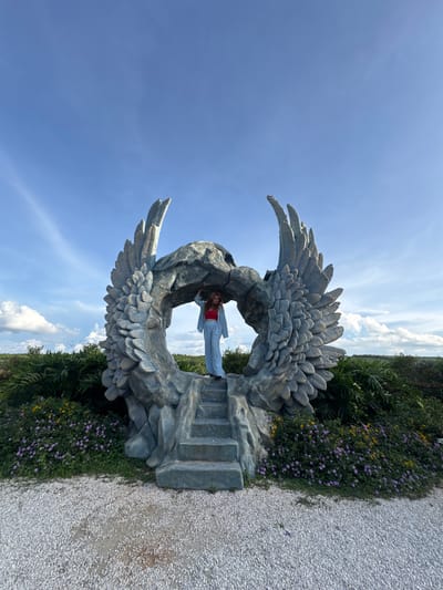 A woman stands on a stone staircase leading into a large, gray, rock sculpture resembling a pair of angel wings arching over a cave-like opening. She wears a red top and light blue pantsuit. The sculpture is surrounded by lush greenery and small purple and yellow flowers. In the background, a vast expanse of blue sky with scattered white clouds stretches above a landscape of rolling hills. The scene appears to be outdoors in a park or garden setting, likely in Thôn 1, Vietnam, given the context of the request. The ground in the foreground is covered in white gravel.