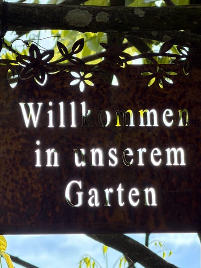 A vertical garden sign in German, "Willkommen in unserem Garten," (Welcome to our Garden) hangs from a tree. The sign is rusted metal with flower-shaped cutouts on its left side. The words are cut out of the metal to allow light to shine through. The sign is set against a natural background of trees and sky. This is located in the city of Esch-sur-Alzette, Luxembourg.