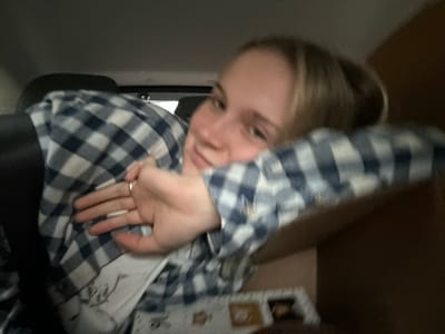 A woman with blonde hair wears a blue and white checkered shirt. She's holding her hand up, partially obscuring her face. She seems to be in a car, with a dark seat visible and the interior of the vehicle in the background. A document or photograph is visible at the top left.