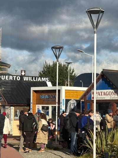 The image displays an outdoor street scene in Ushuaia, Argentina, situated in a commercial area near a waterfront or port, as indicated by "PUERTO WILLIAMS" and "SEABOAT NAVIGATION" signage. Approximately 15-20 individuals, primarily adults, are gathered in small groups, standing on paved and gravel surfaces. They are dressed in warm, casual clothing including jackets, beanies, and backpacks, suggesting a cooler climate. One individual walks on reddish pavers, while a child in pink footwear is seated on a step.
The background features several single-story commercial buildings. One building has a dark corrugated roof with a sign reading "PUERTO WILLIAMS" and an arrow pointing downwards, below which "PINGUINERA" is partially visible. An adjacent building features a wooden facade with "SEABOAT NAVIGATION" text and a poster depicting a boat. To the right, another building exhibits a blue corrugated metal facade and a sign that reads "PATAGONIA Adventure Explore" with a lighthouse graphic.
Multiple streetlights with white poles and dark, inverted-cone-shaped fixtures are distributed throughout the scene. A large green tree is visible behind the buildings, and low green shrubs with small white flowers occupy the immediate foreground. Dark, treeline-covered mountains stand in the distance under a partially cloudy sky with interspersed blue patches. The scene is illuminated by daylight, casting discernible shadows.