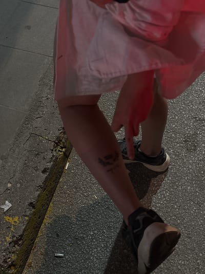 A person's lower legs and feet are visible, standing on a paved surface that appears to be a sidewalk or street. The individual has a tattoo on their left calf. They are wearing dark athletic shoes. The background shows a textured gray surface, possibly concrete or asphalt, with some yellow markings and debris. The lighting suggests it is evening or nighttime. The scene is captured from a high angle looking down. There is no specific indication of the location being Asenovgrad, Bulgaria.