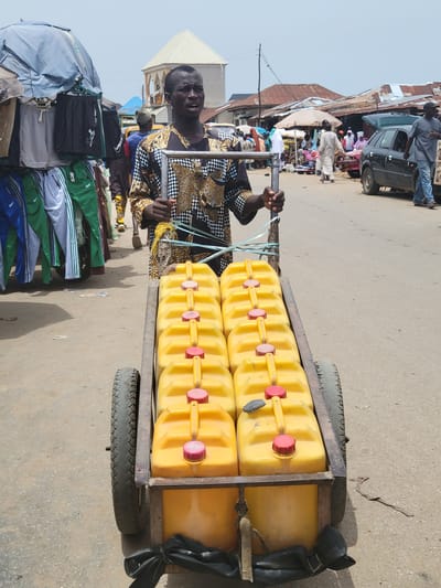 In Suleja, Nigeria, a man stands on a paved street, pulling a wheeled cart loaded with ten yellow plastic jerrycans. The cart, made of metal, has two tires and an improvised braking system consisting of a black plastic sack. The man is wearing a patterned shirt and holds the cart's handle with both hands. Behind him is a display of clothing, including tracksuits, and a blue umbrella. In the background, there are buildings, a black car, and other people. The lighting suggests it is daytime.