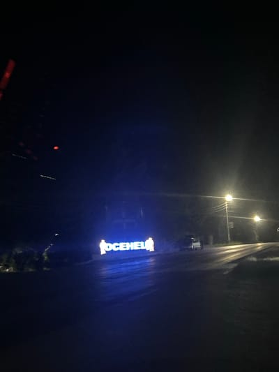 The image depicts a nighttime scene in the city of Osenets, Bulgaria. In the foreground, a sign illuminated with blue neon lights displays the word "OCENETS" with two silhouetted figures standing on either side of the text. The sign appears to be a landmark or an entrance to a facility. The road in front of the sign is wet, reflecting the neon glow and the lights from street lamps. To the right of the sign, a white vehicle is parked, and further down the road, street lights illuminate the path. The background is dark, suggesting it is a rural or suburban area outside of the main city center. The overall atmosphere is quiet and still, characteristic of a night in a smaller Bulgarian town.
