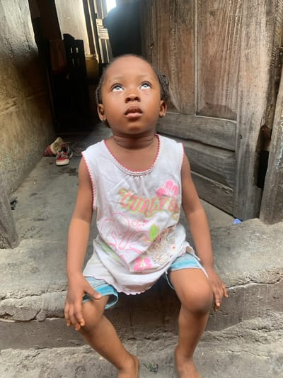 An image shows a young child seated on a concrete step in Okene, Nigeria. The child, with dark skin and braided hair, is looking upwards, with their eyes rolled back to reveal prominent sclera and their mouth slightly open. They are wearing a white sleeveless top with pink and green graphic designs and light blue shorts; they are barefoot. To the child's left, a dark, narrow passage extends into the background, where a pair of red and white sneakers and a metal bowl are visible on the ground. To the child's right, a weathered, vertically planked wooden door stands partially visible. The immediate environment consists of rough, possibly distressed concrete surfaces and aged wooden structures.