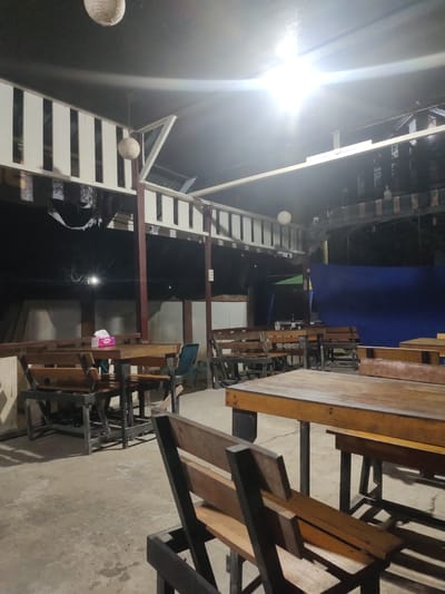 This is a photograph of an outdoor seating area at night, likely a restaurant or cafe, in Keudematangkuli, Indonesia. The scene is illuminated by artificial lights, creating shadows and a slightly dim atmosphere. In the foreground, several wooden tables and benches are arranged on a concrete floor. The tables are simple, with wooden tops and dark metal legs. Some of the benches are also visible, with wooden slats.
In the middle ground, more tables and benches are visible, some with a darker wood tone. A tissue box is placed on one of the tables. In the background, a large blue tarp hangs on the right side, suggesting a temporary or enclosed space. Above, the roof structure is visible, with white slatted railings and dark ceiling panels. Two round, woven light fixtures hang from the ceiling. The overall impression is a casual, open-air dining or gathering space. There are no people visible in the photograph.
