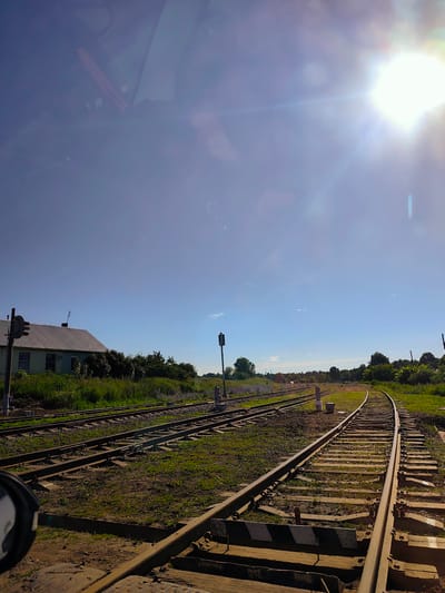 The image depicts a sunny day with a clear blue sky above a rural landscape. In the foreground, a set of train tracks runs through grassy terrain. To the left, a simple, light-colored building with a corrugated metal roof is visible, bordered by dense green foliage. A railway signal with a circular head stands near the building. Further down the tracks, another railway signal with a green light is visible. The tracks curve towards the right, disappearing into a tree-lined horizon. The sun is prominently featured in the upper right corner, casting a bright glare and lens flares across the sky. The scene appears to be a train station or a junction in a rural area of Кварса, Russia, characterized by its natural surroundings and railway infrastructure. There are no people or animals present in the image.
