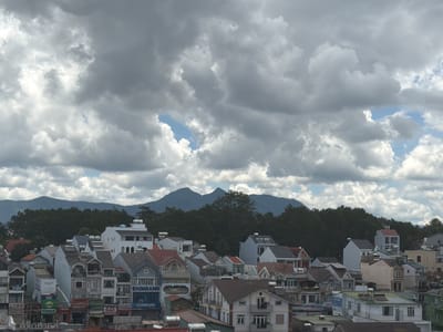 The image shows a densely populated urban area with a mix of architectural styles, characterized by multi-story buildings, many of which appear to be residential homes or small businesses. The structures feature varied rooflines and colors, with a prevalence of red, grey, and white exteriors. Several buildings display signage, including one prominently featuring the name "EXIMBANK" in blue lettering, indicating commercial establishments. Rooftop water heaters are visible on some of the buildings.
Behind the urban sprawl, a dense forest of dark green trees forms a natural backdrop, with two prominent, dark blue-hued mountains rising in the distance. The sky above is filled with dramatic, heavy grey clouds interspersed with patches of bright blue sky, suggesting a dynamic weather system.
The scene is located in Lâm Viên - Đà Lạt, Vietnam, a region known for its hilly terrain, pine forests, and temperate climate, which is reflected in the mountainous landscape and the cloud formations. The architecture suggests a tropical or subtropical influence, common in Southeast Asian cities. There are no people visible in the image, and the focus is on the built environment and the natural landscape.