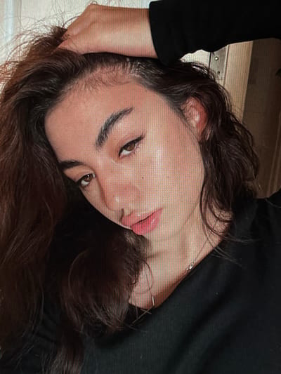 A close-up selfie of a young woman with dark hair, touching her head with her left hand. She is wearing a black top. Her head is tilted, and she is looking at the camera with brown eyes. Her lips are slightly parted, revealing a pinkish hue. The background is blurred, with hints of wood grain and a metal fixture, suggesting an indoor setting. The lighting highlights her facial features, creating a subtle sheen on her skin.