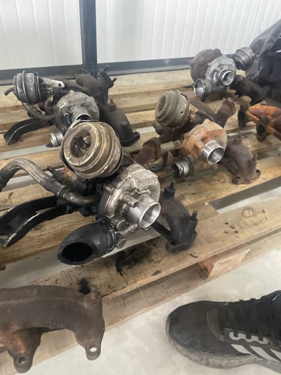 The image captures a collection of used automotive parts, primarily turbochargers and exhaust manifolds, displayed on wooden pallets or beams in what appears to be an indoor setting in Krumovo, Bulgaria.
Several turbocharger units of varying sizes and conditions are visible. Most are metallic, showing signs of wear, dirt, rust, and discoloration, indicating they are salvaged or used components. Some turbochargers include attached wastegate actuators and portions of their associated piping. Interspersed with these are numerous rusty, cast-iron exhaust manifolds, often with multiple ports.
The parts are arranged on rough wooden beams or disassembled pallet sections, which are themselves worn and stained. In the upper right corner, a portion of a person's left foot is visible, wearing a black athletic shoe with white stripes and the number "3" on its side, indicative of an Adidas sneaker.
The background on the left features a white corrugated metal or plastic wall, separated from the foreground by a vertical black metal beam or frame. The floor beneath the wooden structures is a light-colored, possibly concrete, surface. The overall scene suggests a storage area, workshop, or salvage yard for automotive components. No specific activity is taking place other than the static display of these parts.