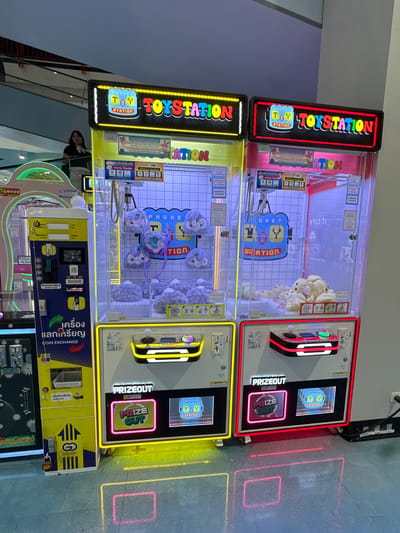 A wide shot captures two brightly lit "Toy Station" claw machines positioned side-by-side in an indoor commercial space, likely a shopping mall, in Wichit, Thailand. The machines are adorned with colorful signage and illuminated displays showcasing plush toys within their glass enclosures. The left machine is predominantly yellow, while the right is pink, both featuring prominent "TOY STATION" logos.

The machine on the left has a "Coin Exchange" section with a basket and instructions. The machine on the right has a "Prize Out" display. The floor beneath the machines reflects the neon lights, creating geometric patterns of yellow and pink. A person, a young woman wearing a dark top and white scarf, is visible in the background, standing on an elevated walkway and looking towards the machines. The overall environment appears clean and well-lit, typical of a retail or entertainment venue.