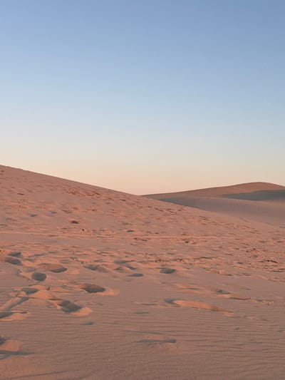 The image depicts a sandy landscape, likely sand dunes, under a clear sky with a gradient from light blue at the top to a pale peach color near the horizon. The foreground and midground are dominated by sand, showing ripples and numerous footprints. In the distance, the landscape curves into rolling sand dunes, casting soft shadows. The lighting suggests it is either sunrise or sunset, with the warm tones illuminating the sand. The location is the Dune du Pilat in La Teste-de-Buch, France, a prominent natural landmark known for its vast sand dunes.