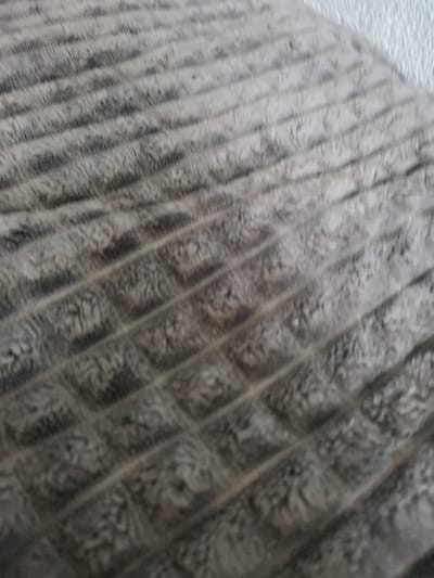 The image displays a close-up, slightly out-of-focus view of a grey, textured textile surface, located in Pokrovnik, Bulgaria. The primary subject is a plush fabric, possibly a blanket or cushion cover, distinguished by a repeating pattern of raised, roughly diagonal linear elements that create a grid-like structure. Within the recessed areas formed by this grid, the fabric exhibits a dense, shaggy, or piled texture, contrasting with the flatter, more defined appearance of the raised lines. The color palette consists of various shades of mottled medium to dark grey, with natural shadows and highlights accentuating the textile's three-dimensional quality. A lighter, subtly textured surface is visible along the upper right edge, indicating an adjacent object or background element. No individuals, specific actions, or other identifying objects are present in the frame.