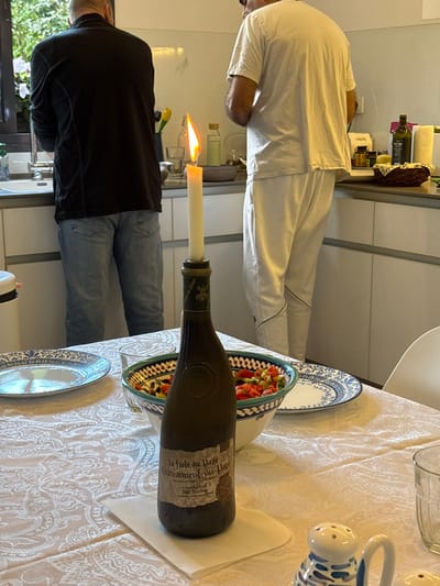 This image captures an indoor scene within a domestic setting in Ramat HaSharon, Israel, depicting a kitchen and an adjacent dining area. In the background, two adult males are visible from the back. The individual on the left wears a dark long-sleeved shirt and blue jeans, standing at a stainless steel sink under a window. The individual on the right wears a white t-shirt and white pants, standing at a kitchen counter with white cabinets below. The counter holds various kitchen items, including an olive oil bottle and a woven basket.
In the midground, a dining table covered with a white patterned tablecloth is set. Centered on the table is a dark green glass wine bottle, identified by its label as "La fiole du Pape Chateauneuf-du-Pape Pere Anselme," repurposed as a candlestick holder. A tall, white tapered candle with a bright, active flame is inserted into the bottle's neck. To the left of the bottle, a white ceramic plate with a blue decorative rim is present. To the right, a similar plate is partially visible, next to a white and blue patterned bowl containing a mixed salad of chopped tomatoes and other vegetables. A clear drinking glass is visible on the far right of the table, and a white and blue patterned salt and pepper shaker set is in the lower right foreground. A portion of a white chair is also visible to the right.