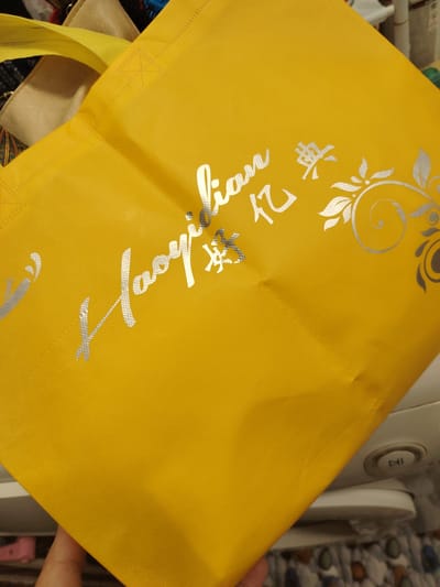 The image shows a close-up view of a yellow fabric bag with silver lettering and a decorative floral design. The lettering appears to be in a script font and is partially visible. The bag is held by a person's hand, which is visible in the lower left corner of the image. The background is blurred and appears to be indoors, with hints of other objects and possibly furniture. There is no indication of the city of Chirchik, Uzbekistan, in the image or its context.