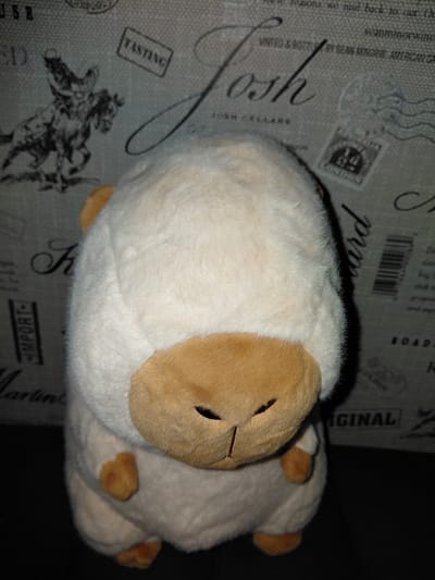 A stuffed toy sheep is positioned centrally in the frame, facing forward. The sheep is predominantly white with tan-colored accents on its face and feet. The background is a textured surface with a repeating pattern of vintage-style graphics, including text, stamps, and illustrations of a cowboy on a horse. The lighting is dim, casting shadows on the right side of the sheep and the background. There is no indication of the city of Vladivostok, Russia.