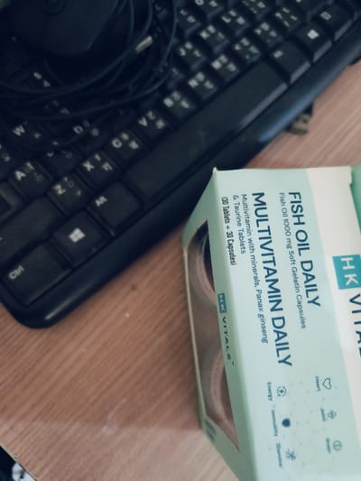 The image shows a black computer keyboard with a mouse lying on top of it, partially visible. The keyboard is on a wooden surface, and a box of "Fish Oil Daily Multivitamin Daily" supplements is partially visible on the right side of the image. The box is light green and white and has the words "Fish Oil 1000 mg Soft Gelatin Capsules" and "Multivitamin with minerals, Panax Ginseng & Taurine Tablets" printed on it. The box also contains a small packet of supplements inside. The coordinates 28.624, 77.055 place the location of the image in New Delhi, India. The image is taken from a slightly elevated angle, looking down at the keyboard and box. The scene is indoors and appears to be a home or office.