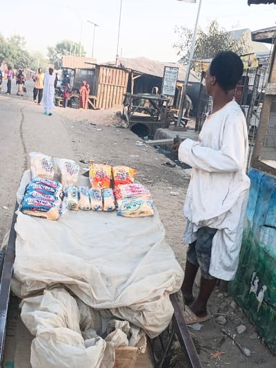 This media file depicts an outdoor street scene in Gawuna, Nigeria, likely a roadside market or vendor stall during daylight hours. The sky appears clear with soft, even lighting, suggesting either morning or late afternoon.
In the foreground to the right, a person stands facing left, looking towards the street. They are wearing a long, light-colored tunic over dark shorts and have short, dark, curly hair. Their right hand holds several small, rectangular items, possibly cards or currency. Immediately to their left is a cart or table covered with a light-colored cloth, displaying various packaged goods for sale. These items, enclosed in clear plastic, appear to be food products like bread or snacks. Visible branding on some packages includes "A.R.M," "SADAQAH," and "SAMIRA."
Behind the vendor, a dusty or unpaved road stretches into the mid-distance, where several people are walking. One person in the mid-left wears a long white garment similar to the vendor's tunic. Along the right side of the road, a narrow open drainage channel runs past several informal structures. These makeshift buildings or stalls are constructed from wood and corrugated metal sheets. A sign on one of these background structures is partially visible, displaying text like "SADAKAN ILLAH" and "MAKA," possibly indicating a religious or charitable message. Further to the right, a simple painting of a black and white cow is visible on a wall or barrier. The scene captures a moment of daily commerce and life in a local community setting.
