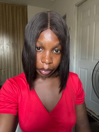 A medium shot captures a woman with dark hair parted in the middle, wearing a red top with a deep V-neck. She has a nose piercing and her skin appears oily. The background includes a white door and a fan partially visible above her. The location is possibly Lugbe, Nigeria. The image appears to be a selfie or taken at close range.