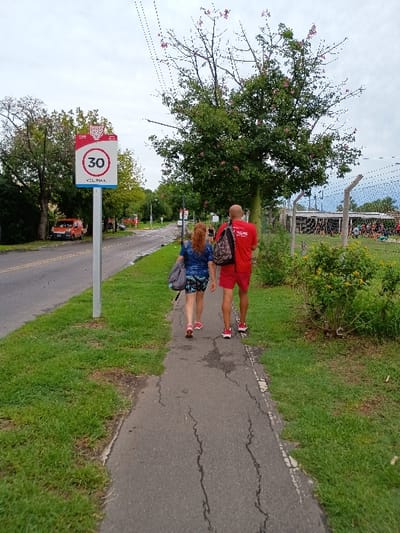 The image displays an outdoor scene in General Pacheco, Argentina, during what appears to be an overcast daytime. Two individuals are captured from behind, walking on a paved pedestrian path that runs parallel to a paved road.
The person in the lead, an adult male, is wearing a red t-shirt with white text (likely "TIGRE") and red shorts, and is carrying a dark backpack. Following behind is an adult female, dressed in a patterned skirt or shorts and a top, carrying a light-colored bag.
To the left of the path, a prominent speed limit sign is visible. It's a white circular sign with a red border, displaying the number "30" in its center. Below the "30", the text "VEL. MAX." (Velocidad Máxima) is clearly readable, indicating the maximum speed. The upper red portion of the sign also features text, possibly "VOS VIVIS TIGRE" or a similar municipal branding, which aligns with the location context of Tigre, a district near General Pacheco.
The path itself shows some surface cracks, suggesting wear. It is bordered by vibrant green grass and various trees. A large tree with distinctive pinkish-red blossoms or berries stands prominently beside the path in the mid-ground. In the background, more trees, some fencing, and other structures are visible beneath a bright, cloudy sky. The setting suggests a public recreational area or a roadside green space.