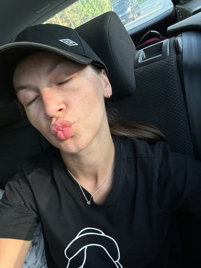 The image shows a person sleeping in a car. They are wearing a black baseball cap with a white logo, a black long-sleeved shirt with a white design, and a silver necklace. The person's head is resting against the car seat's headrest. The interior of the car is visible, including the seat, headrest, window, and seatbelt. The background shows a glimpse of foliage, likely outside the car window.
There is no indication of the location as Langwedel, Germany, within the image. No element definitively places the scene there.