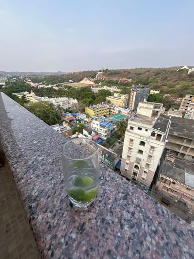 Here is a description of the image:
The photograph is taken from a high-angle perspective, likely from a balcony or rooftop in Puttaparthi, India. In the foreground, a glass of water with slices of lime sits on a stone ledge. The ledge has a mottled dark gray and pink granite-like finish. Beyond the ledge, there is a sprawling cityscape consisting of a mix of low-rise buildings, primarily in yellow, white, and blue. A large hill, covered in greenery, rises in the background to the left, with some buildings nestled into the hillside. The sky is overcast and hazy. In the background, various buildings are visible, ranging from residential to commercial structures. The scene appears to be shot in daylight, although the light is diffused due to the overcast sky. An auto rickshaw sits in front of the buildings in the background.