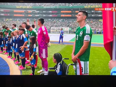 The image shows a televised broadcast of a sporting event, likely soccer, taking place in a stadium. In the foreground, several individuals are lined up on a grass field. On the right, a male athlete wearing a green soccer jersey with white stripes and the number 3 is prominently featured, looking to the right. To his left, another male athlete in a pink goalkeeper jersey stands, facing forward. Several children in matching blue and white soccer uniforms are lined up behind them, some holding hands. A young girl with braided hair is seated in a wheelchair to the left of the pink-jerseyed player. In the background, a large crowd fills the stadium seating, and a scoreboard or banner with the text "MEXICO CITY" is visible. A red flag is held by an unseen person on the far right. The broadcast includes a small overlay in the top right corner with Bulgarian text and a timer, indicating "НА ЖИВО" (Live) and a time of "21:56". The location is not Berkovitsa, Bulgaria, but rather Mexico City, as indicated on the banner.