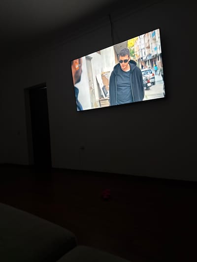 In a dimly lit room, a large flat-screen television displays a scene from a TV show. The show features a young man wearing sunglasses and a black hooded jacket over a dark t-shirt, standing on a city street with buildings and a car in the background. The room itself is sparsely furnished, with a dark wooden floor and a partially visible doorway leading to a darker space. A red object, possibly a toy, is on the floor in front of the television. The overall impression is that of a private residence, likely in the city of Serebryakovka, Russia, where someone is watching television in the evening.