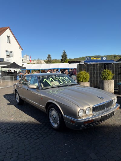 A sideways-oriented, full outdoor shot shows a late-model, light-brown Jaguar sedan in front of a crowd of people gathered under a tent, in Meisenheim, Germany. The car is parked on a cobblestone street. In the background is a building with a sign for "Winters Lebensmittel & Getränke." The sky is clear and blue. A building with a sign and windows is on the lower-left. Another structure, possibly a temporary building or vendor stand, is behind the car, closer to the center. The camera angle suggests the image was taken at ground level. The composition is such that the sedan is a prominent feature, set against the backdrop of a bustling, outdoor gathering.
