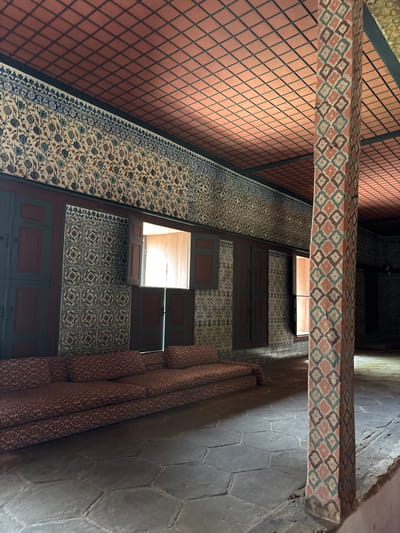 This media file shows an interior view of a hallway with patterned tiles and carpets. The hallway is lit by natural light from an opening above. The walls are tiled with a diamond pattern in a reddish-brown color on the left side, and with intricate blue and white floral patterns on the right side. A patterned carpet with a white background and blue, red, and green floral motifs runs along the hallway. There is a decorative beam overhead with a repeating diamond pattern in red, blue, and white. The floor is made of large, grey hexagonal tiles. The scene is quiet and appears to be within a historical building, likely in Istanbul, Turkey, given the architectural style and tile work. There is no visible text or people in the image, and the time of day is suggested by the light coming from above, which appears to be daytime. The overall impression is one of ornate decoration and historical architecture.