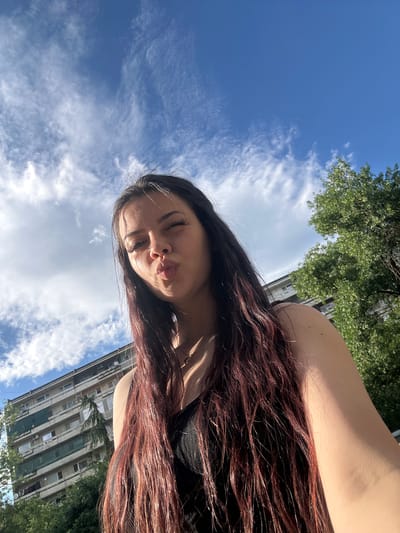 A young woman is depicted in a low-angle selfie, positioned in the foreground and looking upwards toward the camera. She has long, dark hair with reddish-brown highlights, appearing damp or styled, cascading over her left shoulder and chest. She is wearing a dark, possibly black, strapless garment. Her lips are pursed in a kissing or pouting expression, and her eyes are partially closed, with prominent long eyelashes visible. Sunlight illuminates her right shoulder and parts of her hair.
The background reveals an urban outdoor setting in Sliven, Bulgaria. A multi-story residential building, constructed of concrete and featuring numerous balconies, is visible. Some balconies are partially obscured by green screens or tarpaulins. Dense green foliage from trees partially frames the building on both the left and right sides, with a distinct slender conifer tree positioned in front of the building's central section. The sky above is clear and blue, sparsely populated with white cumulus and wispy cirrus clouds. The scene is lit by natural daylight.