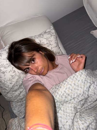A woman is lying in bed in a room with light gray walls and bedding. She is wearing a pink and white striped top. She has brown hair with bangs and brown eyes. She is looking at the camera with a neutral expression. Her right arm is raised and extended towards the camera with a pink bracelet on her wrist. Her left arm is bent. The location is uncertain, and cannot be determined to be Cannes, France.