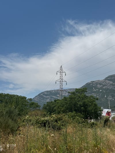 The image depicts a power line tower in a mountainous and vegetated outdoor setting. The tower is constructed of metal lattice and supports multiple power lines that stretch across the frame. Behind the tower, a steep, rocky mountain rises, covered in patches of vegetation. To the right of the mountain, a dense line of trees forms a boundary, beyond which is an area of dry, tall grass and smaller shrubs.

In the sky, wispy white clouds are scattered across a bright blue expanse. The lighting suggests it is daytime, likely mid-morning or afternoon, with clear weather conditions. The overall scene conveys a sense of natural landscape with man-made infrastructure.

In the upper right corner, partially obscured by trees, a billboard with text and an image is visible. The text on the billboard includes "KREDI" which appears to be a word in Cyrillic or Latin script, possibly advertising a financial service or product. A portion of an image of a beverage with a red hue is also visible on the billboard.

The orientation of the image is rotated 90 degrees counter-clockwise. The location context places this scene in Sutomore, Montenegro. No people or specific activities are depicted.
